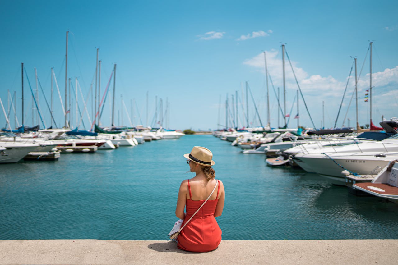 A woman in a red dress and hat sits by a marina with yachts on a sunny day.